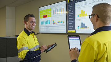 Deux employés de la gestion des installations en conversation, tenant une tablette avec des écrans montrant des données en arrière-plan